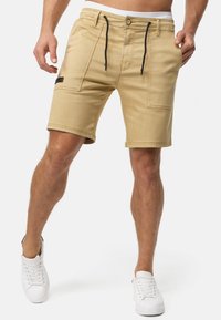 Beige traperice kratke hlače s uskim krojem, koje imaju dvije prednje džepove, pojas s vezicom i oznaku na bočnom rubu. Model nosi bijele tenisice.