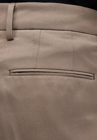 Detailní záběr béžových kalhot z tkaniny, zobrazující zadní kapsu s poutkem na opasek a černý vršek zastrčený dovnitř.