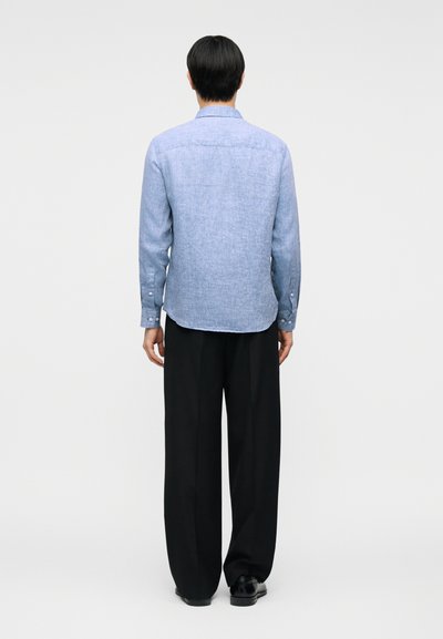 Άνδρας που στέκεται με την πλάτη προς τα εμπρός, φοράει ανοιχτό μπλε πουκάμισο μακρυμάνικο, μαύρο παντελόνι φαρδιάς γραμμής και μαύρα παπούτσια σε απλό φόντο.