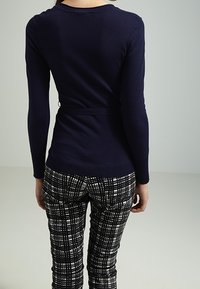 Haut à manches longues bleu marine avec détail cintré à la taille, associé à un pantalon à motif à carreaux noir et blanc. Texture lisse, design simple.