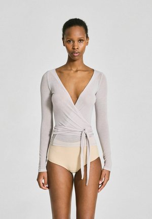 Vrouw staand naar voren gericht, draagt een lichtgrijze wikkelstijl top met lange mouwen en beige hoge taille ondergoed tegen een effen achtergrond.
