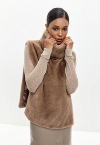 Beige měkká fleece vesta s vysokým límcem; vrstvená přes světle béžový texturovaný top s dlouhým rukávem. Hladká, přiléhavá béžová sukně.