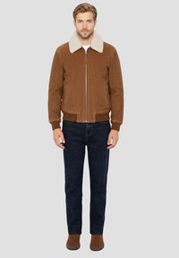 Veste en velours côtelé marron avec col en mouton retourné, fermeture éclair et poignets côtelés, associée à un jean bleu foncé et des chaussures en daim marron.