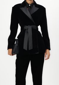 Czarna aksamitna marynarka z satynowymi klapami, z paskiem w talii i długimi rękawami. Modelka trzyma małą czarną torebkę, nosząc czarne spodnie.