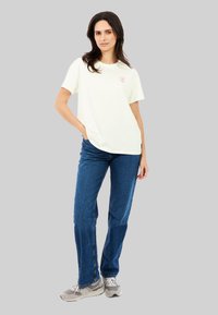 Lichtcreme kleurig T-shirt met korte mouwen en een klein roze grafisch ontwerp op de voorkant, gecombineerd met blauwe jeans en grijze sportschoenen.