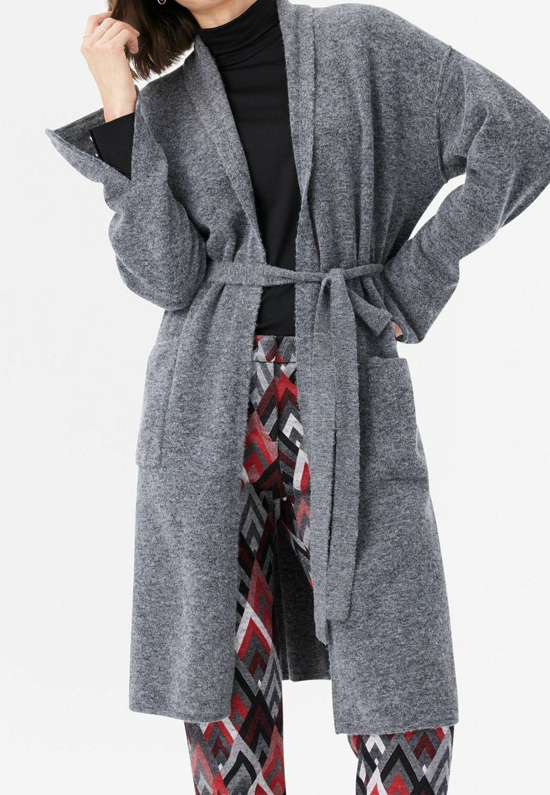 Femme portant un long cardigan gris ceinturé par-dessus un haut noir et un pantalon à motifs rouges, noirs et gris, posant une main sur la hanche.