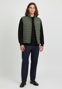 Green quilted vest with a high collar, worn over a black ribbed sweater. Paired with navy trousers and black loafers. Simple logo detail on vest.