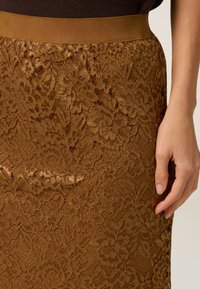 Brun spetskjol med blommönster, som har en mjuk midjeband och en intrikat spetsstruktur. Handen vilar på kjolens sida.