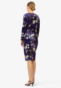 Fluwelen jurk in diep paars met bloemenpatroon, lange mouwen, aangesloten taille en lengte tot de knieën. Heeft een zachte textuur en een elegante uitstraling.