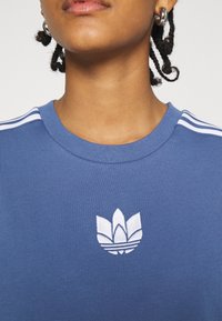 Blå bomullströja med rund halslinning, vit broderad logotyp och tre vita ränder på varje axel.