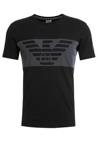 Musta puuvillainen t-paita, jossa on harmaa graafinen kuvio, jossa on kotkaembleemi ja tekstiä. Pyöreä pääntie, lyhyet hihat ja suora helma.