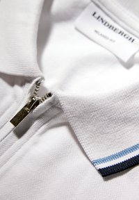 Witte polo met textuur en metalen rits gedeeltelijk open, kraag met zwart-wit-blauwe gestreepte randdetail.