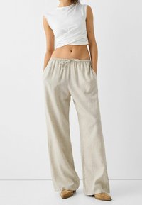 Světlé béžové kalhoty s širokými nohavicemi, elastickým pasem a šňůrkou. Nosí se s přiléhavým bílým topem bez rukávů. Hladký textilní povrch.