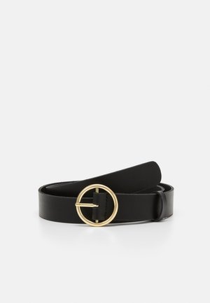 Ceinture en cuir noir enroulée avec une grande boucle ronde en or sur un fond blanc.