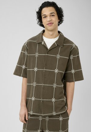 Jeune homme aux cheveux bouclés portant une chemise marron à manches courtes boutonnée et un pantalon assorti avec des motifs blancs en grille et floraux.