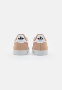 Zapatillas de ante rosa con una suela de goma blanca, que presentan una lengüeta en el talón blanca y un detalle de logo negro en la parte trasera. Diseño minimalista.