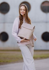 Beige kožený batoh s klopou, nastavitelnými popruhy a embosovaným logem. Měkká textura, obdélníkový tvar a minimalistický design.