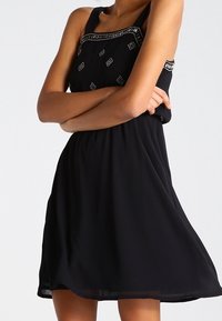 Robe noire à fines bretelles, ornée de détails brodés autour du décolleté. Tissu fluide avec une silhouette à hauteur du genou.