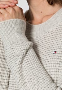 Lichtgrijze gebreide trui met een gestructureerd patroon, geribbelde boorden en een klein geborduurd logo. Zachte stof met een gezellige afwerking.