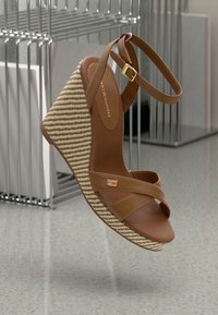 Sandale compensée brune avec des brides entrecroisées et un talon tressé rayé. Présente des garnitures dorées et un petit logo accentué. Texture lisse.