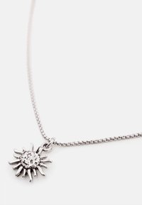 Pendentif argenté en forme de soleil avec un centre texturé suspendu à une fine chaîne en argent sur un fond blanc uni.