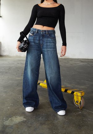 Osoba oblečená v černém crop topu s dlouhým rukávem, modré džínsy s širokými nohavicemi, bílé tenisky a drží malou černou kabelku, stojící na betonové podlaze.