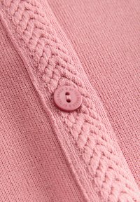 Lichtroze trui met een gemêleerde voorkantknoopsluiting met een gebreide vlecht en een ronde roze knop met het merknaam erop gedrukt.