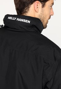 Fekete vízhatlan dzseki magas gallérral és állítható fűzővel. A hátsó részen fehér "Helly Hansen" logó található. Simább felület.