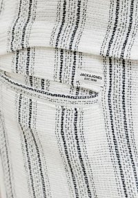 Tissu blanc texturé à rayures verticales noires, avec une poche et une étiquette indiquant "JACK & JONES EST. 1990" sur un vêtement.