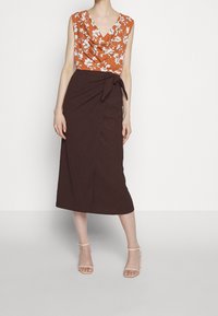 Orange blommönstrad topp med draperad halsringning, parat med en brun omslagkjol med sidoknytning och matchande sandaler med remmar.