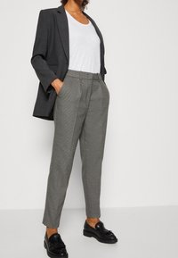 Blazer gris avec un t-shirt blanc, pantalon gris texturé à motif, et mocassins noirs avec des détails en franges. Coupe ajustée et jambes droites.