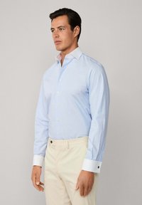 Uomo che indossa una camicia da uomo azzurra chiara con polsini bianchi abbinata a pantaloni beige, in piedi su uno sfondo grigio chiaro uniforme.