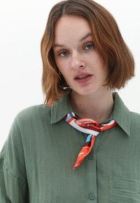 Camicia button-up verde oliva con un motivo testurizzato e una tasca sul petto, abbinata a una sciarpa in seta colorata con accenti rossi, blu e bianchi.