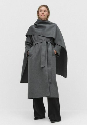 Grå ullinwrap coat med hög krage, bälte och två sidfickor; har en draperad design och strukturerad finish. Kombinerad med svarta byxor.