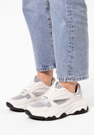 Cream-colored sneaker with mesh and synthetic overlay, black sole, lace closure, and a chunky design. Worn with light blue cropped jeans.