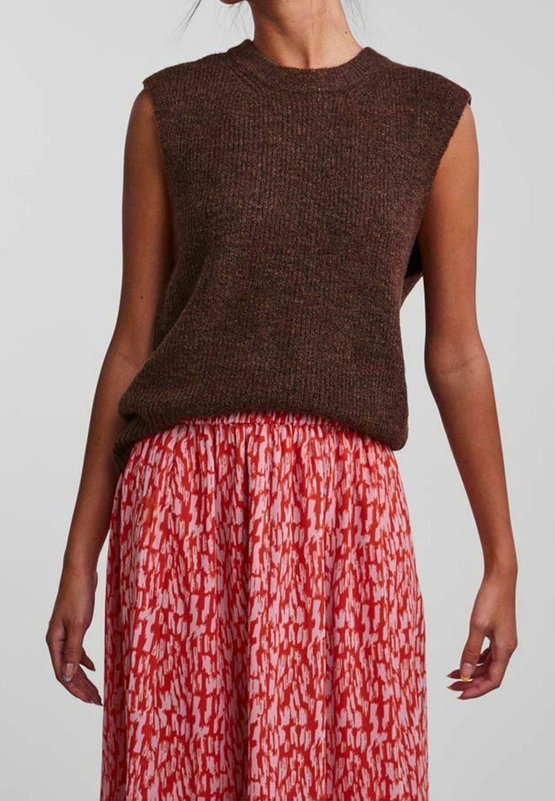 Brun ärmlös stickad topp med ribbat mönster, i kombination med en lång röd kjol med ett vitt abstrakt mönster.