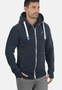 Sudadera con cremallera de forro polar azul marino con cordones blancos, bolsillos laterales y puños acanalados. Presenta un pequeño logo en el pecho.