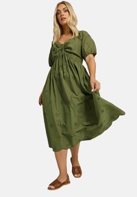 Olivi vihreä mekko, jossa on puffihihat, nauhalla eteen solmittava yksityiskohta ja kukkakuvioisia silkkivaatteita. Helma on aaltoileva ja se on yhdistetty ruskeisiin punottuihin sandaaleihin.