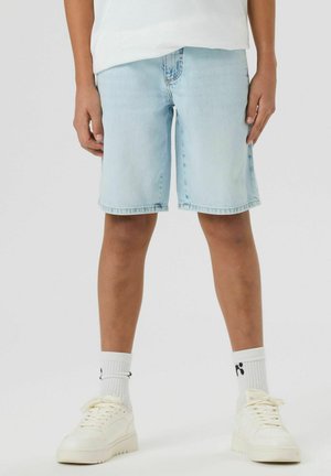 Młoda osoba ubrana w jasnoniebieskie szorty dżinsowe, białe skarpetki za kostkę z czarnym logo oraz białe trampki, stojąca na jednolitym tle.