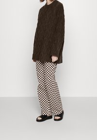 Brauner, texturierter, übergroßer Pullover, karierte, hoch taillierte Hose in Beige und Schwarz sowie schwarze Riemchensandalen werden zusammen getragen.