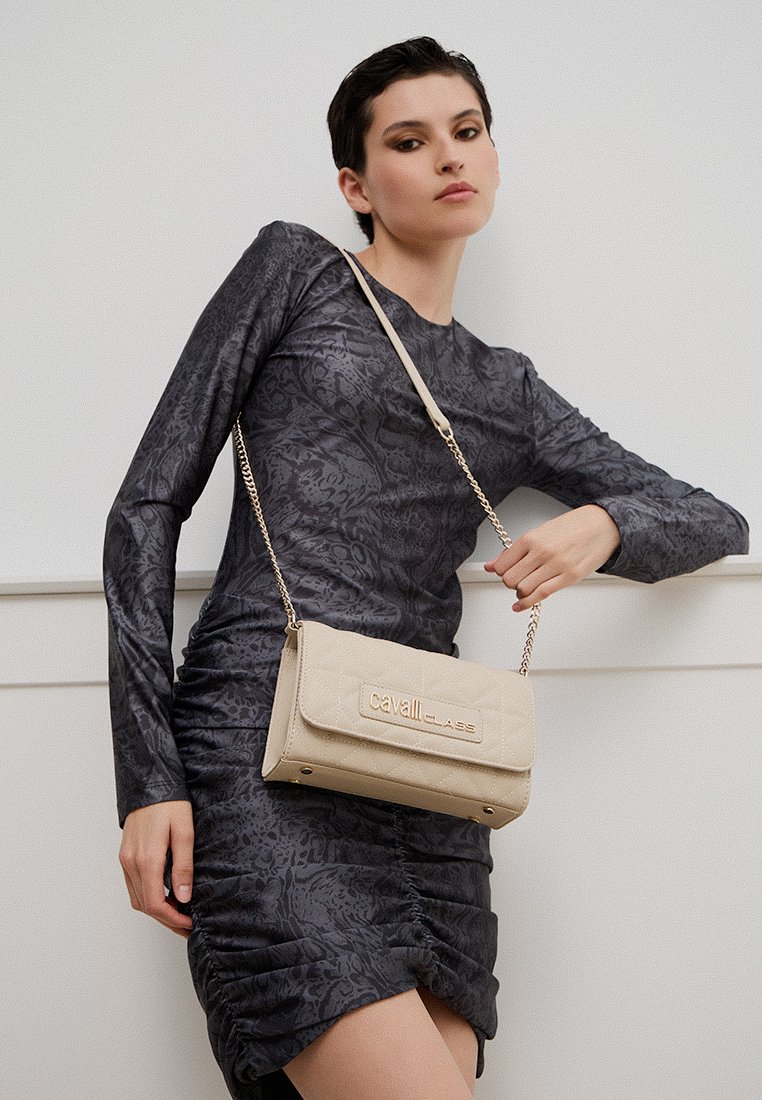 Bolso de mano beige acolchado con herrajes dorados, que presenta un cierre con solapa y una correa de cadena. La modelo lleva un vestido de manga larga con un patrón oscuro.