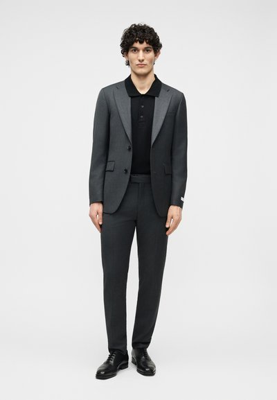 Jeune homme aux cheveux bouclés portant un costume gris foncé, un polo noir et des chaussures de ville noires, debout devant un fond clair uni.