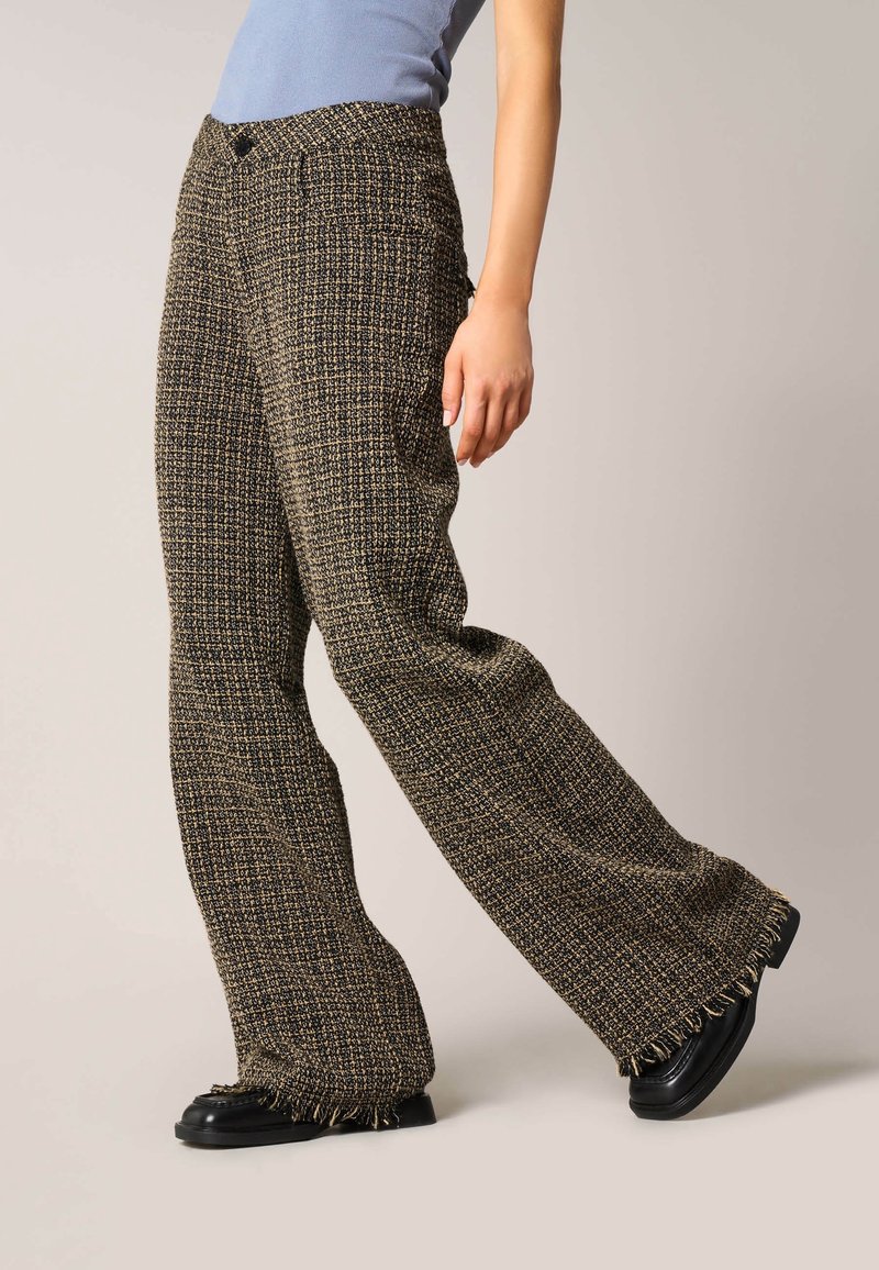Personne portant un pantalon large à carreaux marron et noir et des chaussures noires, posant devant un fond uni clair.