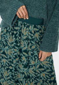 Strikket grøn trøje med brede ærmer, parret med en let grøn nederdel med et blomstermønster i blå og orange, detaljeret med plisseringer.