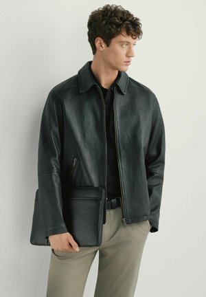 Mężczyzna w czarnej skórzanej kurtce i beżowych spodniach trzyma czarne skórzane portfolio, patrząc na bok na tle jasnego, jednolitego tła.