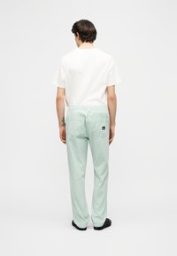 Νέος άνδρας στέκεται με την πλάτη, φοράει λευκό κοντομάνικο πουκάμισο και ανοιχτό πράσινο παντελόνι με πίσω τσέπες και μαύρα παπούτσια.