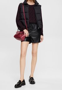 Schwarze Steppweste über einer roten Bluse mit Polka-Dots, kombiniert mit schwarzen Leder-Shorts und glänzenden schwarzen Loafers. Hält eine rote Handtasche.