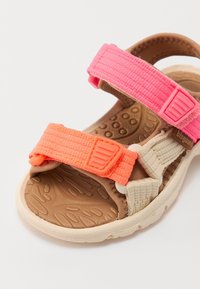 Monivärinen sandaali, jossa on beige kumipohja ja kolme säädettävää remmiä kirkkaan vaaleanpunaisista, oransseista ja kermaista kudottuja materiaaleista.