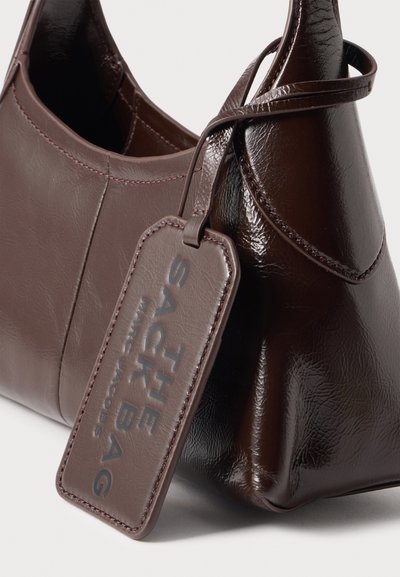 Sac à main en cuir marron foncé avec une forme arrondie et une étiquette assortie embossée avec "THE SHABAG MARC JACOBS."