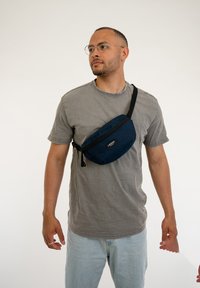 Marsupio blu navy realizzato in tessuto resistente, dotato di una tasca frontale con zip e di una tracolla nera regolabile, indossato a tracolla sopra una maglietta grigia.